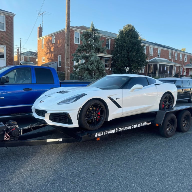 White Corvette being transported on a flatbed tow truck in an urban residential area with a blue pickup truck nearby
