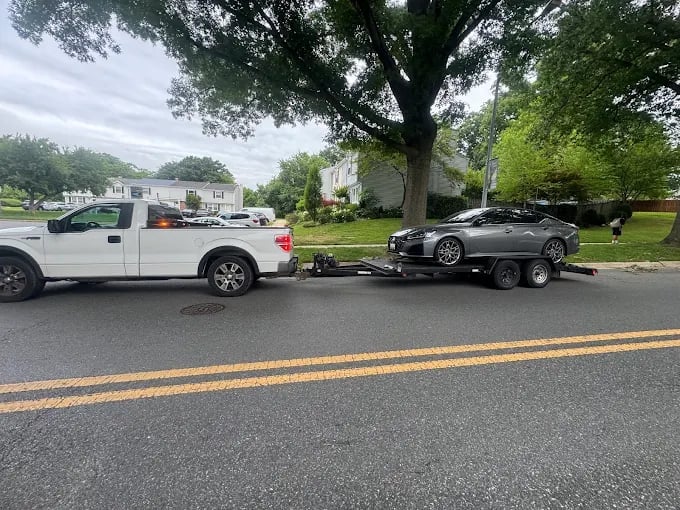 White pickup truck towing a black sports car on a trailer on a residential street lined with trees