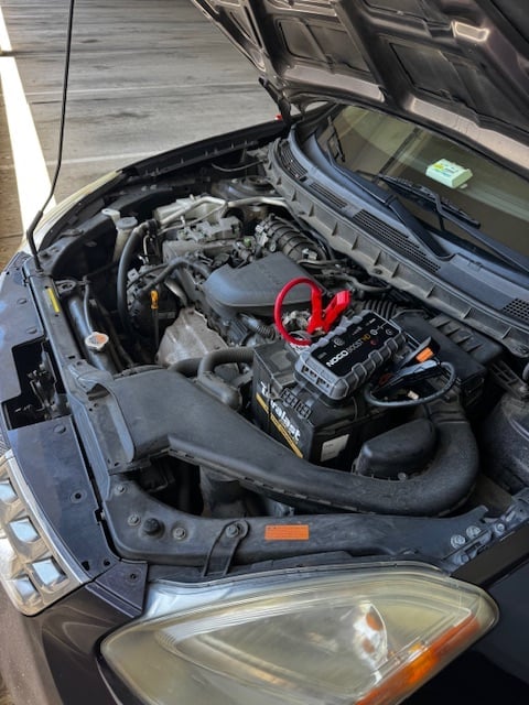 Car engine bay with open hood showing motor, battery with red jumper cables, and black engine components - Jump Start Service Montgomery Village MD