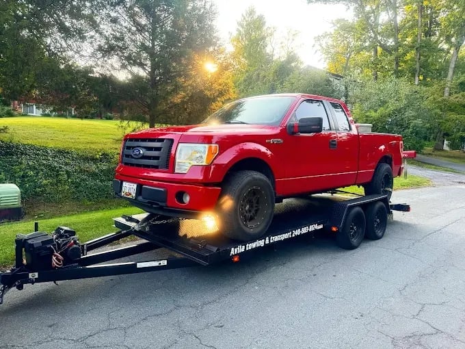 Red Ford pickup truck being transported on a black enclosed trailer in a residential driveway with green trees in background