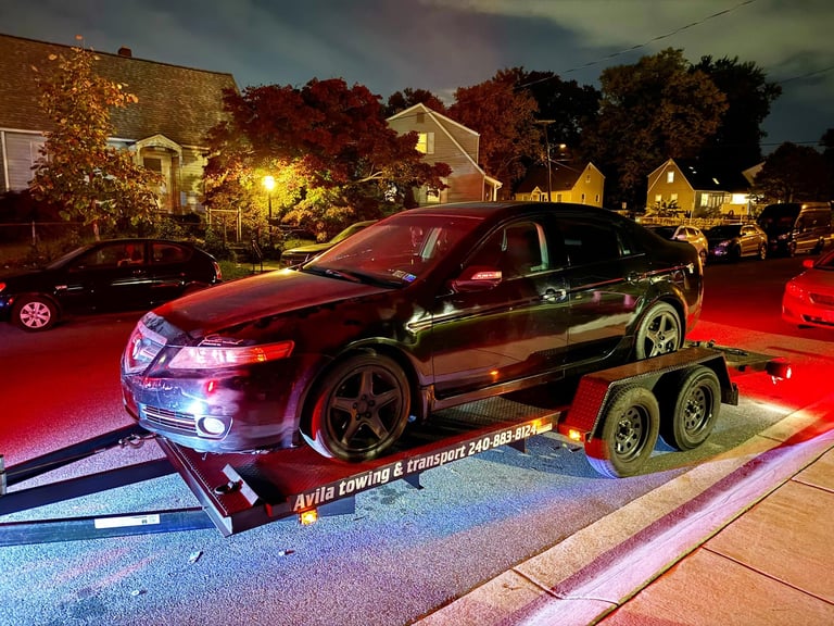 Red car on a two-axle transport trailer in a parking lot at night, with illuminated houses and vehicles in the background