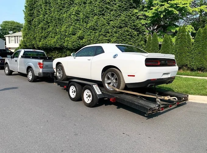 White Dodge Challenger being towed on a flatbed trailer behind a white pickup truck in a residential driveway