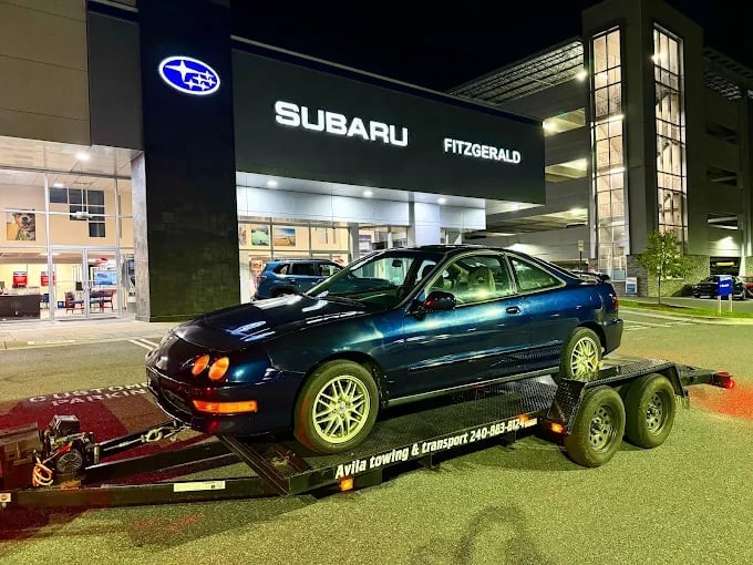 Dark blue sedan on a red tow trailer parked outside a Subaru Fitzgerald dealership at night