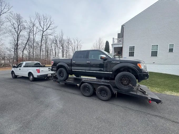 Tow truck hauling a black pickup truck on a residential driveway with trees and white house visible